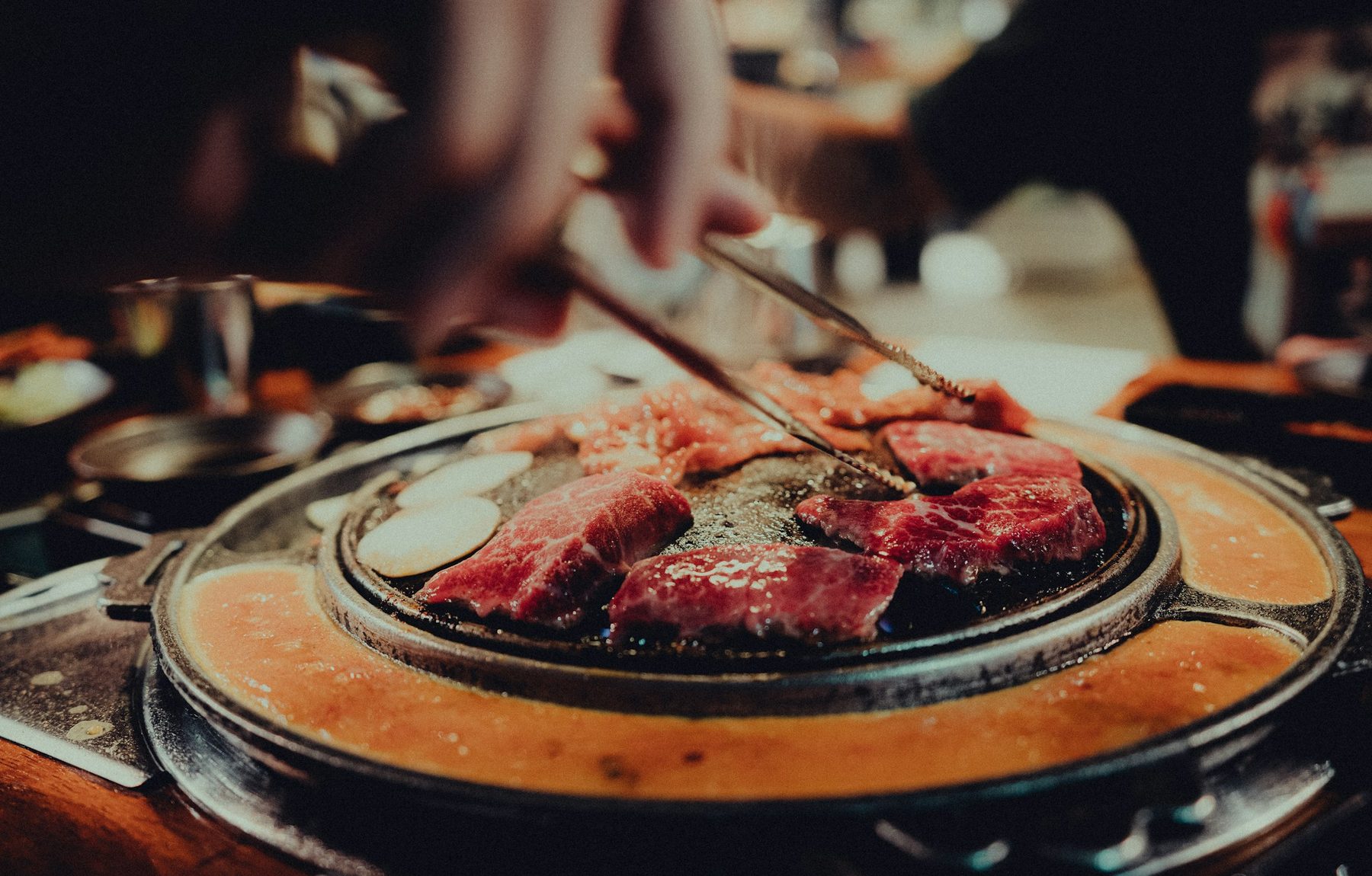 Beef cooking table-side on a Korean BBQ iron plate, tongs turning marbled cuts over the grill